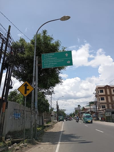 The image shows a street in Katapang, Indonesia (latitude: -6.993, longitude: 107.556) with a street sign pointing to various locations, including Sukamenak, Tol Kopo, Leuwi Panjang, KTR Desa Pangauaban, Rancamanyar, and Cibaduyut. There are power lines strung across the street, and several trees line the edge of the road. A street lamp is positioned on the left side of the frame. There are several vehicles traveling on the road in both directions, including a white van, a green van, motorcycles, and a white car. There is also a yellow sign on the left-hand side of the road that indicates that there is a fork in the road ahead. The sky is blue with white puffy clouds.