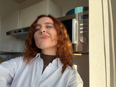 This image depicts a female individual inside a kitchen located in Reims, France. The subject, a woman with auburn, wavy hair, occupies the central foreground. She is dressed in a brown turtleneck beneath an open white button-down shirt. Her head is angled slightly to her right, with her lips pursed in a prominent 'kissing' expression and her right eye partially closed. Her left hand is visible in the bottom-left corner. The background reveals a functional kitchen interior: white overhead cabinets and a stainless steel range hood are visible on the left. On the right, a silver microwave oven with a digital display showing '00:00' rests on a shelf. A turquoise baking dish is partially visible atop the microwave. The scene is strongly illuminated by direct sunlight entering from the right, creating sharp shadows and highlighting a textured cream-colored wall.