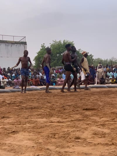 The image shows a wrestling match taking place on a sandy ground in Sokoto, Nigeria. The location is (latitude: 13.049, longitude: 5.238). There are several men in the wrestling ring, with at least five men standing in the ring. The men are mostly shirtless and wearing shorts. One man is wearing a white shirt and a white hat. Many people are sitting on the ground and watching the wrestling match. They are wearing traditional clothing. The wrestling match is taking place in a large, open area. There are buildings in the background. The sky is gray and cloudy. The men in the ring are about to wrestle. They are looking at each other, and some of them are making gestures. There is a small crowd of spectators watching the match. They are sitting on the ground, and some of them are talking and laughing.