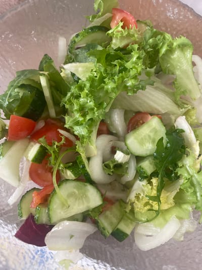 This media file shows a close-up, overhead view of a fresh salad in a clear bowl. The salad contains various ingredients, including frilly green lettuce, sliced cucumbers, halved cherry tomatoes, and thinly sliced white onion rings. Some arugula leaves are also visible, adding a dark green contrast. The vegetables appear crisp and vibrant, suggesting they are recently prepared. The lighting suggests it could be daytime, though the specific time is not determinable. The setting appears to be indoors, possibly a kitchen or dining area, given the salad's presentation in a bowl. No people, text, or specific location cues are evident within the image itself, beyond the general context of a prepared food item. The overall impression is of a healthy and appetizing meal.