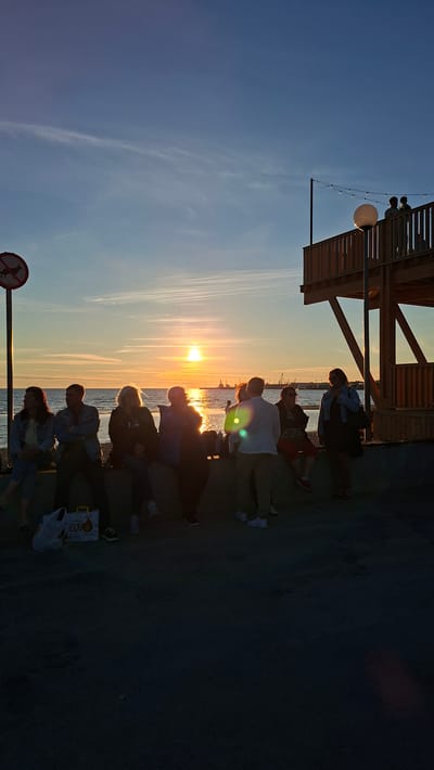 This is a vertical, eye-level, full shot of a group of people gathered outdoors by a body of water at sunset. The sun is low in the sky, casting a warm, orange glow across the scene and reflecting on the water. The sky is a gradient of blue and orange, with some wispy clouds.

On the left side of the image, a signpost with a red and white "no dogs allowed" symbol is visible. To the right of the sign, several people are sitting on a low wall, facing the water and the sunset. From left to right, the first person is a woman wearing a light-colored jacket and pants. Next to her is a man in a dark jacket. Then, a woman with blonde hair wearing a dark jacket. Following her is a woman in a blue jacket. The last person on the wall appears to be a woman in a dark top.

In front of the people sitting on the wall, there are two bags on the ground, one white and one yellow.

To the right of the sitting group, more people are standing and interacting. A man in a white shirt and khaki pants is facing away from the camera. To his left, a woman in a dark dress is standing. Further to the right, a wooden structure, possibly a viewing platform or part of a building, rises up. It has railings and is lit by string lights. Two figures are silhouetted against the sky on the upper level of this structure. A large white spherical lantern is also visible on this platform.

The overall atmosphere is relaxed, with people enjoying the sunset. The location appears to be a public space along the waterfront, with a cityscape or industrial area visible in the distance across the water. Given the presence of a sign in Russian and the architectural style of the wooden structure, this scene is likely located in Tallinn, Estonia, specifically in the area of the Linnahall or the nearby harbor.