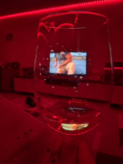 The image shows a wine glass in a room lit with red light, possibly from an LED strip. Inside the glass, a reflection of a phone screen displays an image of a person. The background is blurry, but suggests a bedroom environment with furniture and bedding. The location, Ust-Ilimsk, Russia, cannot be confirmed or denied based on the visual information alone.