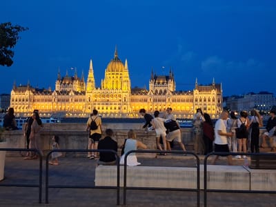 This image captures the Hungarian Parliament Building in Budapest at dusk, illuminated by artificial lights against a deep blue sky. A river, likely the Danube, is visible in the mid-ground, with a boat passing by. In the foreground, a group of people are gathered on a paved promenade overlooking the river and the iconic building.
The individuals are engaged in various activities: some are standing and conversing, others are seated on a low concrete wall. A young child in a light-colored dress is walking away from the camera. The people are dressed in casual summer attire, suggesting a warm evening. The architecture of the Parliament Building is elaborate, featuring numerous spires, domes, and arched windows, all highlighted by a warm, golden glow. The overall atmosphere is serene, with the twilight setting lending a sense of calm to the bustling city scene.