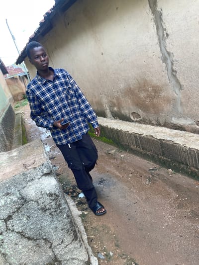 A young man walks through a narrow, damp alleyway with concrete gutters on either side. He is wearing a blue and white plaid long-sleeved shirt, black cargo pants, and black sandals. He holds a dark-colored smartphone in his right hand. The buildings flanking the alley are made of aged, light-colored concrete with weathered roofs. The ground is a mix of mud, gravel, and debris. The scene appears to be located in Rafin Par, Nigeria, as indicated by the context. The overall atmosphere is slightly overcast.