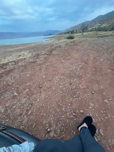 The image shows a dirt ground with small rocks, a body of water and mountains in the background. A person wearing black slippers and dark clothing is sitting near a car. The location is likely near Bin El Ouidane, Morocco, given the landscape. The sky is cloudy.