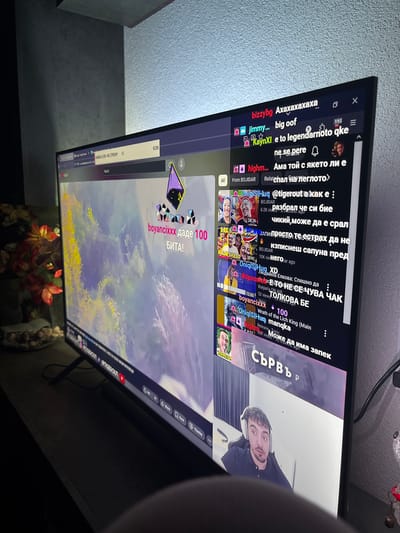 This image, captured in the city of Kuklen, Bulgaria, displays a television or monitor angled slightly to the left, functioning as a display for what appears to be a live YouTube or Twitch stream. The screen exhibits a multi-panel interface: the largest section on the left presents stylized digital artwork resembling a landscape with vibrant yellow, green, and purple hues. Overlaid on this art is a purple and gold geometric icon, associated with Twitch "bits," and the Cyrillic text "boyancixxx даде 100 БИТА!" (boyancixxx gave 100 BITS!). The right side of the screen features a vertical chat window, predominantly displaying messages in Bulgarian. Notable chat entries include "bizzybg Axaxaxaxaxa," "jimmy... big oof," "KaynXI e to legendarnoto qke ne se pere," "highm... Ама той с якето ли е," "OniqlSHuq @tigerout a как е :/", and explicit comments from "BOJIDAR" such as "разбрал че си бие чикий, може да е срал" (understood that he is masturbating, might have shat) and "Запипросто те естрах да не изписнеш сапуна пред него" (Just... you're afraid not to write the soap in front of him). Other messages mention "Людмила Елкова: Спешно да," "tdripsosetd ЕТО НЕ СЕ ЧУВА ЧАК ТОЛКОВА БЕ," "OniqlSHuq Wrath of the Lich King (Main manqka)," and "highm... Може да има запек." The bottom right corner of the screen shows a smaller webcam feed of a male streamer with dark hair and a beard, wearing a dark shirt and white over-ear headphones, looking directly forward with a slightly wide-eyed expression. The top bar of the interface shows a search bar containing "VARNA VLOG + IRL STREAM" and "€150," suggesting a stream title and a monetary goal or total. Partially visible navigation elements and playback controls are present at the top and bottom of the screen, including a YouTube icon. The monitor rests on a dark, flat surface, likely a wooden cabinet or desk. To the left of the monitor, on the same surface, is a decorative bowl filled with light-colored, round objects, possibly pebbles or beads, positioned in front of a vibrant artificial plant with red and orange foliage. The background on the right reveals a light-colored, textured wall. A black power cable descends from behind the monitor, leading off-frame to the right. A small decorative object with red accents is partially visible on the far right. The overall lighting is dim, suggesting an indoor setting illuminated primarily by artificial light, making the screen