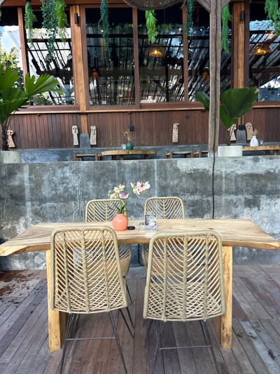 The image captures an outdoor seating area of a restaurant or cafe with a rustic and natural aesthetic. In the foreground, a large, rough-hewn wooden table is set with four woven rattan chairs. The table has a live edge and its surface shows natural wood grain and imperfections. On the table, a small, pale pink and white orchid arrangement in an orange vase is visible, along with a small black object and a small white glass. A menu is also propped up on the table.
The seating area is constructed with natural materials. The floor is made of weathered wooden planks, with some damp patches suggesting recent rain or cleaning. Behind the table and chairs is a rough, textured concrete wall, which provides a backdrop to the scene.
Above the concrete wall, a wooden facade with vertical planks is visible, featuring several decorative wooden human-like sculptures mounted on it. A large window or glass facade with dark wooden framing is set into this wooden structure, reflecting the sky and the interior of the building, which appears to have hanging plants and woven light fixtures.
The overall environment is lush and green, with large palm-like plants positioned to the left and right of the wooden facade. Hanging plants are also present above the wooden structure, adding to the tropical or bohemian atmosphere. The lighting suggests it might be late afternoon or early morning, with soft natural light. The scene appears to be an outdoor dining area designed to blend with its natural surroundings. The location is identified as Kediri, Indonesia, suggesting a tropical climate.