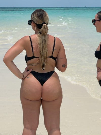 Two women are standing at the water's edge on a sandy beach in Zanzibar, Tanzania, facing the clear, turquoise ocean under bright daylight. The primary subject, positioned centrally, is seen from behind. She has blonde hair tied in a wet ponytail and wears a black two-piece swimsuit consisting of a bikini top and a high-cut thong bottom. She wears dark rectangular sunglasses and has a script tattoo on her lower back, a small butterfly tattoo on her upper left thigh, and a camera outline tattoo on her left upper arm. Her hands are placed on her hips. A second woman is partially visible on the right side of the frame, facing the ocean and wearing a black bikini top and dark sunglasses. The water shows subtle ripples near the shore, transitioning from clear shallows to deeper turquoise. In the far distance on the horizon, a faint dark object, possibly a boat, is discernible.