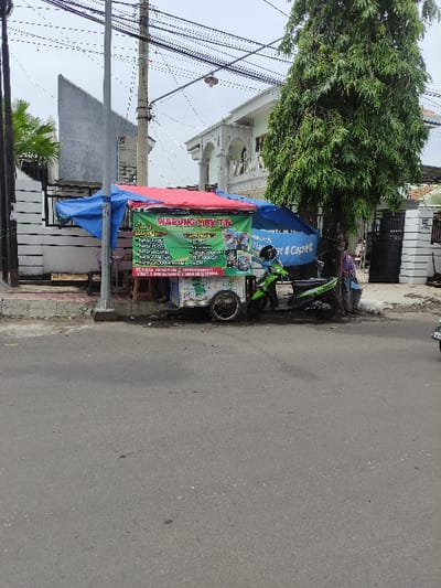 The image captures a daytime street scene in Situbondo, Indonesia, featuring a makeshift street vendor stall alongside a paved road and sidewalk. The lighting is bright and diffused, suggesting either an overcast sky or early morning/late afternoon.

In the foreground, a large utility pole stands on the left, crisscrossed by numerous overhead power lines. To its right, a vendor stall is set up on the sidewalk. The stall is constructed with a table covered by a red tarpaulin canopy, with blue tarpaulins draped down the sides, providing shade and shelter. A green banner on the front of the stall prominently displays the name "MARUKOMEN TRE" and advertises various snacks and drinks. The menu includes "Snack & Minuman" (Snacks & Drinks), "Jasuke" (Corn Milk Cheese), "Cilor" (Tapioca egg snack), "Sempol," "Cilok Kuah" (Tapioca balls with soup), "Mie Goreng" (Fried Noodles), "Indomie Kuah" (Indomie with soup), "Sosis Bakar" (Grilled Sausage), "Kentang Goreng" (French Fries), "Pop Ice," "Es Teh" (Iced Tea), "Es Jeruk" (Orange Juice), and "Kopi" (Coffee). Above this, a partially visible blue banner displays text ending with "... & Cepat" (... & Fast). Adjacent to the food stall, another green and white banner is visible, likely advertising services such as "Jualan Pulsa" (Selling credit/top-up) and "paket data" (data packages).

A green motorcycle is parked next to the vendor stall on the sidewalk. Beyond the stall, a large, dense green tree is prominent. In the background, behind the tree and power lines, various buildings are visible, including a notable white structure with arched windows that resembles a mosque or a large residential building. The asphalt road in the immediate foreground is empty, and no people are visibly engaging in activities, though the presence of the stall implies commercial operation. The scene depicts a common urban or semi-urban street environment in Indonesia.