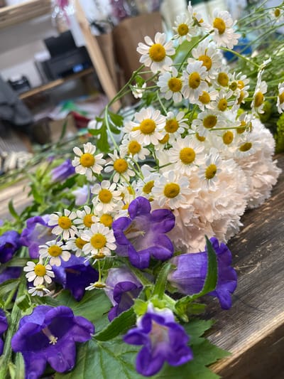 A close-up, high-angle shot shows a bouquet of flowers on a wooden surface. The bouquet is primarily composed of small white daisies with yellow centers and vibrant purple bell-shaped flowers. Interspersed are large, pale pink, ruffled carnations and a hint of green foliage. The background is out of focus but suggests an indoor environment with shelving and possibly some office equipment. The lighting is natural and soft, highlighting the textures and colors of the flowers. The scene appears to be in the city of Kommunarka, Russia, likely in a florist's shop or a home setting.