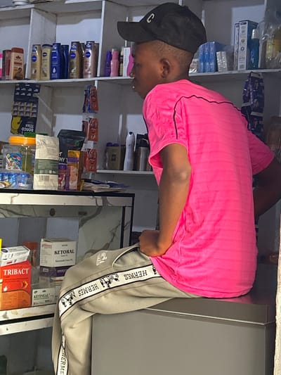 The image depicts the interior of a small shop, likely a pharmacy or convenience store, located in Ihiagwa, Nigeria. A young person wearing a bright pink t-shirt and grey pants with a distinctive "HERMES" logo on the side is seated on a counter. The shelves behind them are stocked with various products, including toiletries, medicines, and personal care items. Some of the visible brands include NIVEA, TRIB ACT, and KETORAL. The setting appears to be dimly lit, with artificial lighting illuminating the shelves. The overall impression is that of a bustling local retail environment.