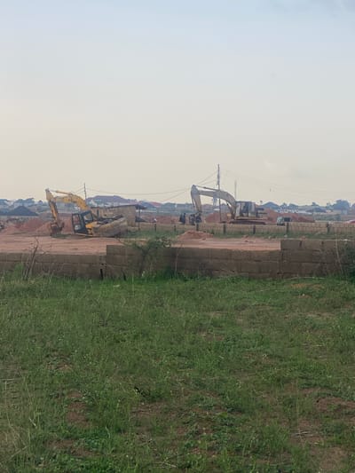 In Kwang, Nigeria, this image captures a construction site with two excavators actively engaged in earthmoving. The foreground is dominated by a low wall constructed from concrete blocks, with a patch of green grass in front of it. In the middle ground, a yellow excavator is positioned on a mound of earth, while another, larger excavator is seen further back, also amidst piles of soil. Several individuals are present, with some gathered near the larger excavator, appearing to be workers or supervisors. The background reveals a residential area with houses and buildings, suggesting the construction is part of a larger development. Power lines crisscross the sky, indicating an urban or semi-urban setting. The overall atmosphere is one of ongoing construction and development within a community.