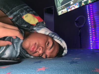 A person is lying beneath a blue blanket with star patterns. The person has dark hair, brown eyes, and appears to be male. He is partially wrapped in a grey and black blanket with yellow and red patches. In the background, there is a television screen displaying various content, a ring light hanging on the wall, and LED strip lights emitting blue and red light. The location, Parroquia Ocumare, Venezuela, cannot be verified from the image.