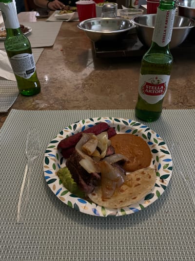 This image captures an indoor dining scene, situated within a private residence or casual restaurant in Monterrey, Mexico. In the foreground, a white paper plate, adorned with a green and blue leaf pattern, holds a prepared meal. This meal consists of slices of cooked meat topped with sautéed white onions, a serving of refried beans, several pieces of red sausage, a portion of guacamole, and one folded flour tortilla. To the left of the plate, on a light green woven placemat, a clear plastic fork is positioned. A green glass bottle of Stella Artois beer, approximately two-thirds full, stands above the plate. Another similar green beer bottle is partially visible to the upper left. The table surface is a light brown, mottled material, possibly granite or marble. In the midground, other dining items are present, including red plastic cups, silver metal bowls, a silver salt shaker, and white dishes. In the background, a person's arm and hands are visible, engaged in an activity at the table.