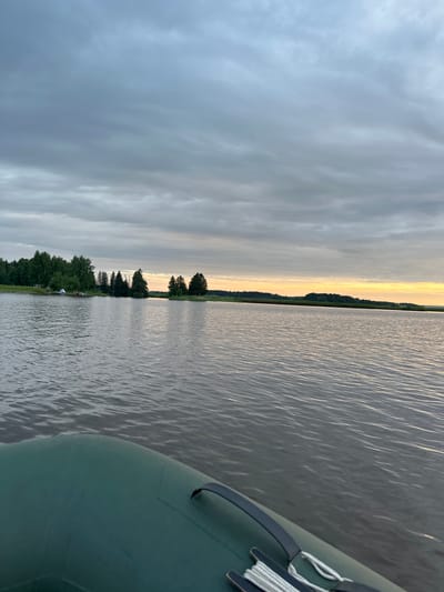 An over-the-shoulder shot from a boat shows a calm lake with a distant tree-lined shore under a cloudy sky. In the foreground, the green inflatable boat has a rope coiled on its deck. The lake water is dark and rippled. The far shore is mostly covered in dense green trees, with a few smaller islands also visible. The sky is overcast with grey clouds, but a sliver of yellow light on the horizon suggests the sun may be setting or rising. The location is Shul'dikha, Russia.