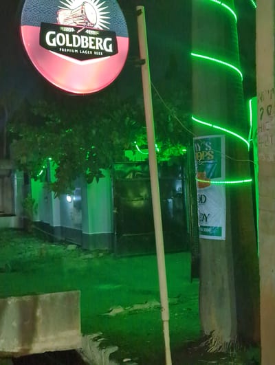 The image shows a nighttime scene in Narayi, Nigeria. A "Goldberg Premium Lager Beer" sign is visible on the left. Green LED lights are strung along the upper portion of a building facade. A "DY TOPS" sign is above a dark doorway. Vegetation, likely a small tree, obscures part of the view. The environment has an overall green hue from the lighting.