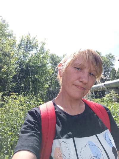 A blonde woman with fair skin and freckles, wearing a black t-shirt with a cartoon print and a red backpack, is captured in a selfie. She is outdoors, with a bright sky and lush green trees in the background. A portion of a grey pipe is visible to her right. The scene suggests a casual, outdoor setting, possibly in the city of Dyurtyuli, Russia.