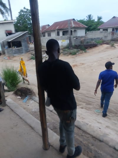 The image displays an outdoor scene in Mwandege, Tanzania, featuring three individuals and various structures along a dirt road under an overcast sky.
In the foreground, a male individual is positioned with his back to the camera, standing next to a prominent vertical wooden pole. He wears a black t-shirt, light-colored long pants (possibly jeans) with a visible dark patch on the right thigh, and dark shoes. A light-colored item, possibly a pouch, hangs from his left hip.
To the right of the foreground individual, on the unpaved dirt road, a second male figure walks away from the camera. He is dressed in a blue t-shirt, blue jeans, and a dark baseball cap. Further to the left and slightly behind the foreground person, a third individual, obscured partially, walks wearing a bright yellow garment that appears to cover most of their body.
The immediate ground consists of a concrete curb running along the lower left side, with green bushy vegetation behind it. The dirt road occupies the center and right, showing an uneven surface with scattered refuse, including a light blue plastic bag.
The background features several buildings under construction or in various states of completion. On the far left, a structure with a corrugated silver roof and light-colored walls is visible. Towards the center and right, multiple buildings with reddish-brown corrugated metal roofs and light-colored concrete walls are present. Some of these structures appear unfinished, displaying exposed concrete and construction debris, including stacked rebar-like materials. A small yellow sign is visible on the ground or a low wall between some of these central background buildings. Palm tree foliage is visible at the top edges of the frame.