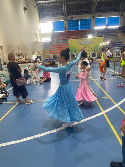 The image captures an indoor event, likely a dance competition, held in a gymnasium in Thuận An City, Vietnam. The floor is a blue gymnasium surface marked with yellow and white lines. In the foreground, a young girl in a light blue dance dress with a full, pleated skirt is captured mid-motion, with her arms extended. To her right, a younger girl in a pink dance dress with a similar full skirt is walking, being guided by an adult. Further back, a woman in a black dress with a purple handbag over her shoulder is standing and observing. Several other individuals, including adults and children, are visible in the background, some sitting on the floor and others standing near a banner that reads "GRACE ACADEMY". The gymnasium features tiered seating in the background, some of which is orange and yellow. Lights hang from the ceiling, illuminating the space. The overall atmosphere suggests a formal event with participants and spectators.
