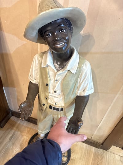 An upward-angled photograph depicts a full-body statue of a young boy positioned indoors in Tangier, Morocco. The statue, appearing to be crafted from painted resin or ceramic, features a dark complexion, a broad smile revealing white teeth, and dark eyes. It is dressed in a light-colored, wide-brimmed hat, a cream-colored collared shirt with rolled sleeves, a light tan vest with four dark buttons, light tan trousers, and a black belt with a silver buckle. Both of the statue's hands are extended forward with palms open and facing upwards.
In the foreground, a person's right hand, wearing a silver band on the ring finger, reaches towards the statue's right hand. The person's arm, clothed in a dark blue jacket sleeve, extends from the lower left of the frame. The background consists of a plain, light beige wall with subtle vertical patterns and a light-colored wooden floor.