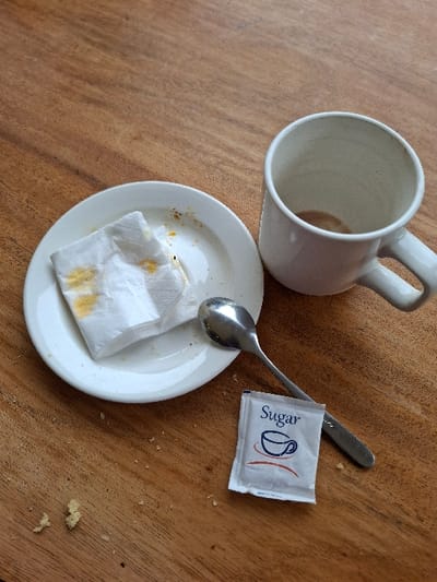 The image shows a white mug, a white saucer with a napkin and a few crumbs on it, a small silver spoon and a white sugar packet on a wooden table. The table appears to be made of medium brown wood with a slightly rough texture. There are no people or animals present. The location is Karagita, Kenya, based on coordinates (latitude: -0.772, longitude: 36.421). The mug is empty, and the spoon is lying on the table beside the saucer. The sugar packet appears to be unopened, and there is a crumb on its surface. The napkin is also partially on the saucer. The scene suggests that someone recently had coffee or tea.