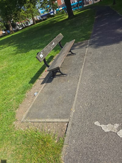 Two park benches, made of dark wood and metal, are situated on a paved area next to a grass lawn. The benches are angled towards each other. The grass lawn is green and well-maintained, with scattered small white flowers. Beyond the lawn, trees with full green foliage are visible, and in the background, buildings and a blue bus are present, suggesting an urban setting within Stockton-on-Tees, United Kingdom. The paved area on the right of the image shows an asphalt surface with some white markings and scattered cigarette butts. The ground next to the benches is dirt and also contains litter, including cigarette butts and a red object. The lighting suggests it is daytime, with shadows cast by the benches and trees.