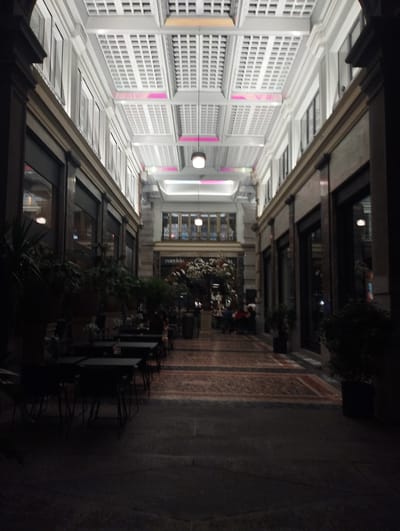 The image captures an interior view of a covered arcade or gallery at night. The architecture features high, vaulted ceilings with a grid-like structure illuminated by soft, indirect lighting, including pink and white glows. On either side of the central walkway are the facades of buildings with large windows, some reflecting interior lights. The walkway itself is paved with a patterned carpet or tile. In the foreground and mid-ground, there are several dark tables and chairs set up, suggesting an outdoor seating area for a restaurant or cafe. Some plants in dark planters are also visible. In the background, more architectural details and lighting are apparent, with hints of people seated at tables further down the arcade. The overall impression is one of an elegant, dimly lit public space at dusk or night. This location appears to be within the GUCCI store on Via Nassa in Lugano, Switzerland, based on the architectural style and the "GUCCI" sign visible in the background.