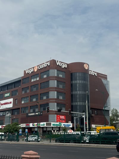 This is a frontal, eye-level medium shot of the Vega Centre, a modern, multi-story commercial building in Tashkent, Uzbekistan. The building is predominantly reddish-brown with large, dark windows. Prominent signage includes "Vega Centre" in white lettering on the upper facade, with a red circular logo. Other visible signs on the building include "EVOS," "korzinka.uz," "TERRA PRO," and various Cyrillic text. A curved glass section is visible on the right side of the building. In the foreground, a street with multiple lanes of traffic is present. Several cars, including a white car with a patterned wrap, are visible on the street and parked behind a green metal fence. Traffic lights are also visible. The sky is overcast. The overall scene depicts a busy urban environment.