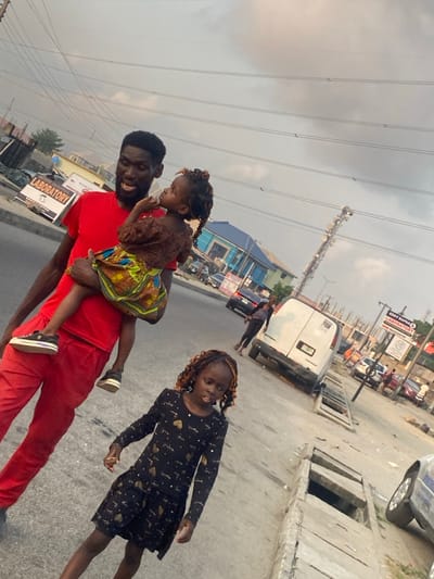 The image, taken in Badore, Nigeria, captures a man and two young girls outdoors on a street or roadside under an overcast sky. The photograph is tilted, giving a dynamic perspective to the scene.
In the foreground, a man with a beard, wearing a red t-shirt and red pants, smiles warmly as he carries a young girl. This girl, with dark braided or twisted hair, is dressed in a brown top and a vibrant, colorful patterned skirt. She looks intently towards the left side of the frame. To their right, a slightly older girl stands, looking towards the viewer or slightly past. She has dark hair with lighter streaks and is wearing a black long-sleeved dress adorned with gold heart-like patterns. Both girls appear to be young children.
The background reveals an urban or suburban setting. Power lines stretch across the grey, cloudy sky, and a tall communication tower stands prominently in the mid-ground. Various buildings line the street, including one with a distinctive blue facade. Vehicles, including a white van, are visible parked or driving further down the road. Concrete drainage covers are present along the road's edge. A sign in the distance is partially readable, displaying "BRODY ALPHA" and "LABORATORY," with "HOSPITAL" also partly visible. The overall scene depicts a casual daytime outing for the man and the children, under a diffused, cloudy light. The man's smiling expression suggests a pleasant moment.