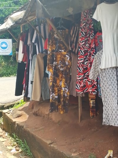 The image depicts a roadside clothing stall in Umuahia, Nigeria. A collection of garments, including dresses and tops, hangs on hangers under a makeshift shelter constructed from poles and a tarp or fabric covering. The stall is positioned on a raised dirt and concrete platform along the roadside, with the road visible in the background alongside a sign for the Redeemed Christian Church of God, Abia Province 5. The ground around the stall is dirt and vegetation.