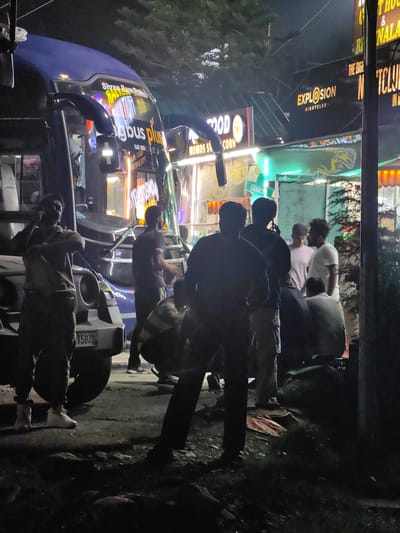 The image depicts a nighttime scene in Sambal, India, featuring a group of people gathered near a large bus and a vehicle. A prominent sign for "BIGBUS PLUS" is visible on the side of the bus, suggesting it is a travel service. Adjacent to the bus, several businesses are illuminated by neon signs, including "EXPLOSION NIGHTCLUB" and a food establishment advertising "MOMOS SPICY CORN." The general environment appears to be a commercial or roadside area with trees present in the background.
In the foreground, several individuals are visible. One person, wearing sunglasses and a short-sleeved shirt, is standing on the left, appearing to be engaged in a conversation or making a gesture. To their right, another person in a dark t-shirt is seen interacting with a group of individuals in the center of the frame. A silver SUV-like vehicle is parked in front of the bus, its license plate partially visible. The ground is asphalt, with some debris and shadows adding to the nighttime atmosphere. The lighting suggests a mix of streetlights and commercial signage.