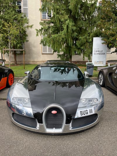 The image displays three Bugatti supercars parked on an asphalt surface, with a prominent chrome and black Bugatti Veyron in the foreground, facing the viewer. This main vehicle features a chrome silver finish on its front fenders and lower bumper, contrasting with a glossy black hood, roof, and central engine cover. A white license plate reading "BUG III" is affixed to the front bumper, below the right headlight. The iconic Bugatti grille with its red oval badge is centered on the front.

To the left, partially visible, is the front-left quarter of another Bugatti, dark-colored (likely black) with bright orange multi-spoke wheels and a subtle rear spoiler. To the right, also partially visible, is the front-right quarter of a third Bugatti, appearing in a metallic dark brown or bronze hue, with a silver multi-spoke wheel.

In the background, behind the vehicles, are several large green trees with dense, weeping foliage. Behind the trees, a light-colored building with multiple windows featuring closed white louvered shutters is visible. To the right, a tall, white vertical sign reads "Lycée CAMILLE SCHNEIDER" in large black lettering, with "DESIGN ALSA" in smaller text below it. This identifies the location as Lycée Camille Schneider in Molsheim, France. Adjacent to the sign is a gray outdoor trash receptacle with a black bag liner. No people are visible in the scene. The overall environment suggests an outdoor parking area, possibly on school grounds.