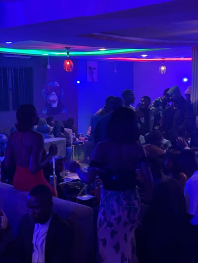 In Durumi, Nigeria, a dimly lit indoor venue, possibly a club or lounge, is captured in the image. The space is illuminated by vibrant neon lights in shades of blue and green, casting a cool glow on the scene. Several people are present, some of whom are dancing or socializing. A woman in a red dress is visible in the foreground, facing away from the camera. Another individual, a man in a suit, is seated on a couch to the left. A prominent mural of a monkey wearing sunglasses adorns one of the walls. The atmosphere appears lively and energetic, typical of a night out.