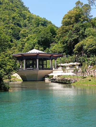 The image shows a tranquil scene at the Abkhazian city of New Athos. A vibrant turquoise lake dominates the foreground, its surface gently rippling under the clear blue sky. In the mid-ground, a distinctive pavilion-like structure stands on a raised platform overlooking the water. The pavilion features intricate dark blue and red detailing around its ornate roof, supported by white classical columns. A black railing encircles the platform.

Adjacent to the pavilion, to the right, is a terrace area with several white umbrellas providing shade for outdoor seating. People can be seen seated at tables or strolling along the terrace, enjoying the view. The architecture of this area is characterized by tiered levels with stone retaining walls and lush greenery. Further to the right, more people are visible on a balcony area, some sitting and others standing.

The background is a steep, verdant hillside densely covered with lush green trees, creating a dramatic natural backdrop. The overall impression is one of a peaceful, picturesque resort or park setting.