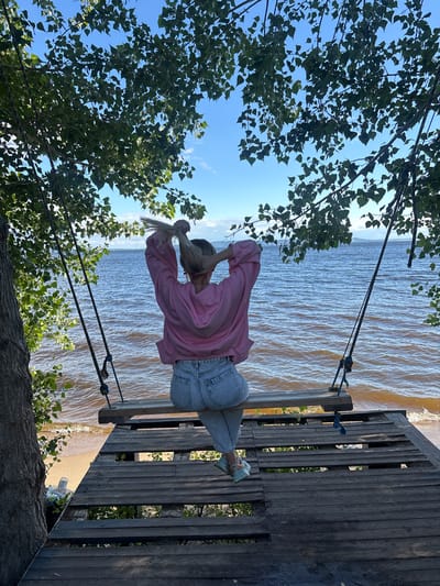 A woman is sitting on a wooden swing overlooking a body of water. She is wearing a pink hoodie and blue jeans. Her back is to the camera, and she is seen tying her blonde hair into a ponytail. The swing is suspended by ropes from a tree with green leaves. The platform of the swing is made of weathered wooden planks. In the background, the water is rippling, and the sky is blue with some clouds. The scene appears to be located in Podstepki, Russia, on a beach near the water.