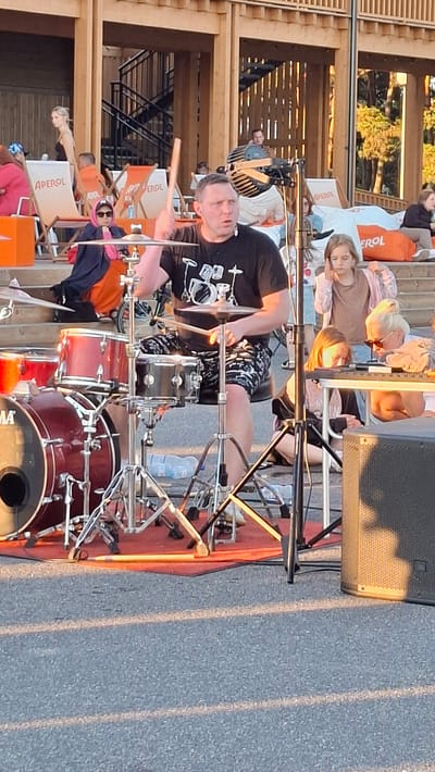 The image shows a man playing drums outdoors during what appears to be a festival or concert. The drummer, a middle-aged man with short hair, is wearing a black t-shirt with a graphic print and patterned shorts. He is actively engaged in playing his drum set, which includes several cymbals and drums, with drumsticks in his hands.

In the background, a wooden structure with horizontal slats suggests an outdoor venue, possibly a stage area or seating section. Several people are visible in the background, some seated on chairs and others standing. The lighting indicates it is either late afternoon or early evening, with the sun casting long shadows on the ground. The weather appears to be pleasant, as people are dressed casually.

The chairs in the background have visible text on them, including the word "APEROL," suggesting a sponsor or a branded event. The overall scene conveys a lively atmosphere, typical of an outdoor music event. The location is identified as Tallinn, Estonia, adding specific geographical context to the scene.