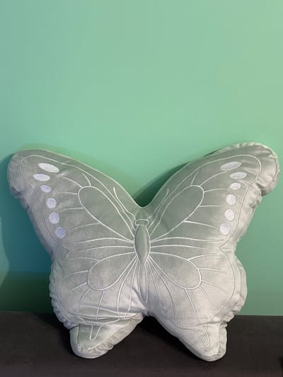 A light mint-green, plush, butterfly-shaped pillow rests on a dark, flat surface. The pillow is centrally positioned in the lower portion of the frame, featuring white embroidered outlines depicting wing veins and a central body, along with vertical rows of white embroidered circular spots on each wing. The background consists of a solid teal-green wall, which occupies the upper two-thirds of the image. A subtle shadow is cast by the pillow onto the wall on its left side. The scene is located in Voluyak, Bulgaria.