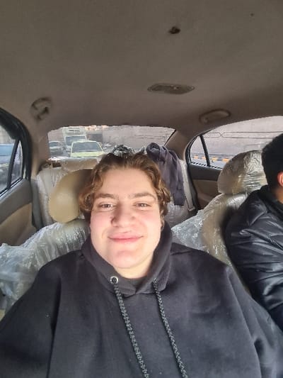 A person with curly brown hair and a black hooded sweatshirt sits in the passenger seat of a vehicle. The interior has light tan upholstery, partially covered in clear plastic. Another person wearing a black jacket is partly visible in the background. The vehicle is on a street with buildings and other cars visible through the window. The photo appears to be taken at an angle, with the interior of the car in the foreground.