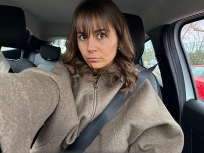 A female subject is depicted in a close-up, front-facing view, seated in the driver's position of a stationary vehicle, within the city of Mougins, France. The individual, appearing to be in her 20s or 30s, possesses medium brown hair styled with bangs and waves. She is dressed in a light brown or taupe zippered jacket/coat and is restrained by a dark-colored seatbelt diagonally across her chest and horizontally across her lap. Her facial expression is neutral, with a slight downturn of the mouth, and her gaze is directed towards the camera. The vehicle's interior features dark-colored headrests and seat upholstery, with a subtle textured pattern visible on the seat to the subject's right. The passenger-side window, visible on the right side of the frame, exhibits water droplets, indicating rain or recent precipitation. Through this window, a red vehicle is partially visible, parked adjacent to the subject's car, along with bare tree branches against an overcast sky, suggesting an outdoor setting such as a parking lot or street.