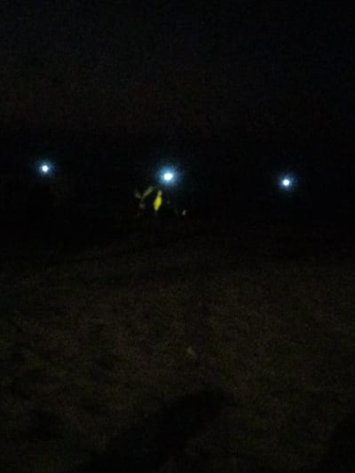 This image depicts a nighttime scene in Al-Mawasi, Palestinian Territories. The environment is extremely dark, with the foreground and midground consisting of indistinct, dark, textured ground, possibly unpaved terrain or sparse vegetation. The sky is completely dark. Three distinct, bright, circular light sources are visible against the dark background. One light is positioned in the upper-left portion of the frame. Another light is in the upper-right portion. A third light source is located near the center of the frame, slightly to the left of the upper-right light. Directly below and slightly to the left of this central light, a blurry, indistinct yellowish-green form is visible. No clear individuals, actions, or other specific objects or interactions are discernible due to the profound darkness and blur.