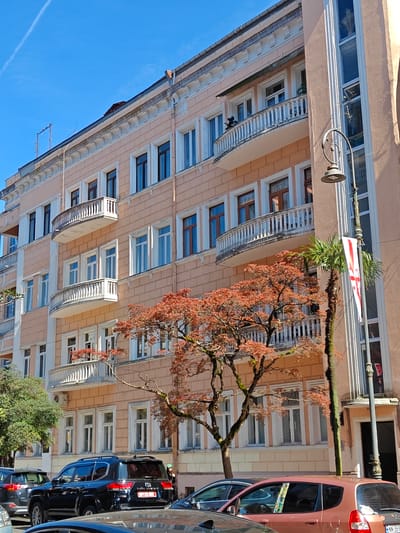 A street view in Batumi, Georgia shows a multi-story peach-colored building with white balconies and dark-framed windows. A tree with red-orange leaves partially obscures the building's facade. A streetlight with a banner displaying the Georgian flag stands near the building. Multiple parked cars occupy the foreground, including a black SUV with a visible license plate. The sky is clear and blue with a contrail in the upper left.
