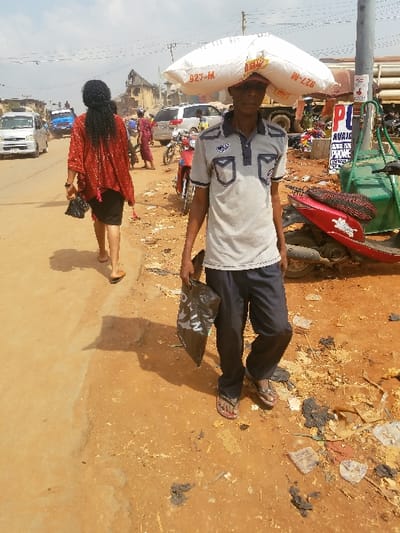 The image depicts an outdoor street scene in the city of Nnobi, Nigeria, under bright daylight. In the foreground, a male individual walks left-to-right, carrying a large white sack labeled "927-M" on his head and a black plastic bag in his left hand. He wears a grey short-sleeved polo shirt with dark blue pockets and collar, dark trousers, and dark flip-flops. To his left, slightly behind, a female individual walks away from the camera, wearing a red patterned top, a dark skirt, and light-colored flip-flops, carrying a small black handbag in her left hand.
The ground is unpaved, consisting of reddish-brown dirt with scattered debris, litter, and dark, irregular clumps. Shadows from the people and objects indicate strong overhead sunlight. In the mid-ground, a paved road is visible, with several vehicles, including a white mini-van and other cars, along with parked motorcycles. A blue vehicle is further down the road. Several other people are present in the background, some walking, some near vehicles. Utility poles with overhead power lines traverse the scene. On the right, a utility pole features a red and white sign with the letters "PU" prominently displayed, and a smaller sign partially visible below it. Various buildings, including a multi-story structure with a distinct roofline, are visible in the background, lining the street.