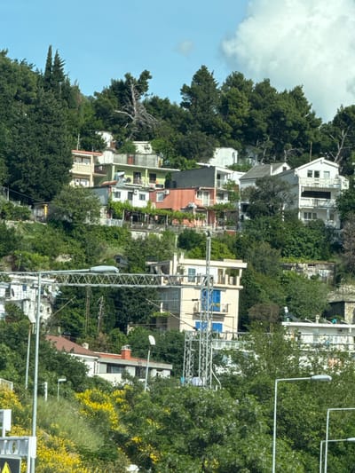 The image shows a hillside in Sutomore, Montenegro, densely populated with houses and trees. The houses vary in color and architecture, interspersed amongst the greenery. The sky is blue with some clouds. Power lines and street lights are visible throughout the scene.