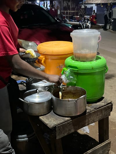 The image captures a bustling scene, likely a street food vendor in Barikin Gidar Kanat, Nigeria. The primary subject is a person wearing a red shirt, with their arm extended as they interact with a food preparation area. They appear to be holding a piece of food, possibly an egg, in a white bowl.
Surrounding the person are various containers and cooking implements. A large, bright green bucket is prominently featured, possibly used for storing ingredients or as a cooler. Next to it, an orange cylindrical container is visible, suggesting it holds more food items. Several metal pots are stacked, with one larger pot on the right appearing to contain some yellowish liquid, hinting at a dish being cooked or served. A transparent plastic container with a pink handle sits on top of the green bucket. The entire setup is placed on a rustic wooden structure, likely a makeshift cart or table, typical of street vendors.
The background shows a hint of a marketplace or a busy street with people and vehicles, including motorcycles, further reinforcing the setting. The lighting suggests it might be evening or night, given the artificial lights visible in the distance and the overall ambiance. The scene conveys a sense of everyday commerce and food preparation. There is no visible text in the image.
