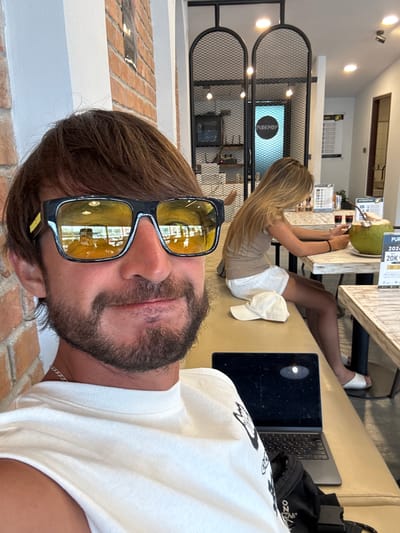 A close-up selfie shows a man with brown hair and a beard wearing sunglasses with yellow lenses. He is wearing a white t-shirt. In the background, a woman with blonde hair is sitting at a table, looking at her phone. She is wearing a beige top and white shorts. A laptop is on the table in front of the man. A green coconut with a straw is on the table in front of the woman. The scene appears to be a cafe in Rawai, Thailand, with exposed brick walls and metal fixtures.