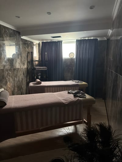 The image depicts two massage tables set up side-by-side in what appears to be a spa or therapy room. The room has marble-patterned walls and dark blue curtains covering a window. Each massage table is covered with a light pink and white striped sheet and has a grey towel folded at the head. One of the tables also has a rolled grey towel at the head and a folded grey towel on top of the sheet. A black shelving unit with towels and small potted plants is visible to the left of the tables. A clock is mounted on the wall above the curtains. The lighting is dim, with shadows cast by the massage tables and some decorative plants in the foreground. The scene is clean and organized, suggesting a professional setting. The location is Hisarya, Bulgaria, a city known for its spa resorts and mineral springs.