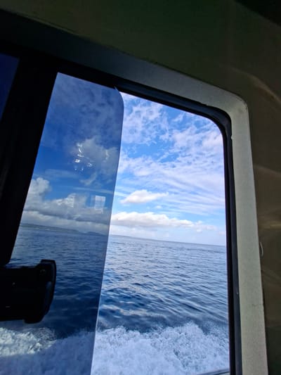A view from inside a boat looking out of a window reveals a bright blue sky with scattered clouds. The ocean stretches out to the horizon, with gentle waves and white foam from the boat's wake visible in the foreground. The window frame is visible on the right side of the image. The boat appears to be moving, as indicated by the wake. Reflections on the glass of the window show the sky and clouds, as well as a dark object, possibly a camera, in the lower left corner. The scene is set on the ocean, with a distant shoreline visible in the background.