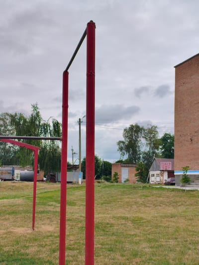 The image captures an outdoor scene on a cloudy day in Ustyvytsia, Ukraine. In the foreground, two tall red metal poles supporting a horizontal bar are prominently featured, with a third red pole visible further to the left. A section of a red frame is also visible on the left. The ground is covered in dry, patchy grass.
In the mid-ground, several structures are present. To the left, there is a truck with a hay bale on it and a large cylindrical tank. Behind these, a red tractor is visible. Further to the right, a small brick building with a gray door and a separate, slightly dilapidated white and red building with a pitched roof and a basketball hoop are situated. A dark-colored car is parked in front of the white and red building. Trees, including a large weeping willow, are scattered throughout the scene, and power lines run overhead, supported by utility poles. A tall, unfinished brick building stands to the far right of the frame, its upper portion obscured by the angle. The sky is overcast with gray clouds.