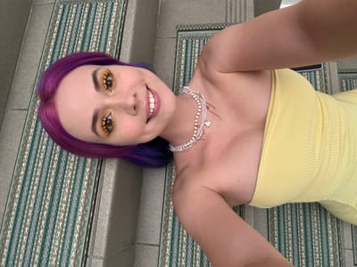 A woman with purple hair and yellow makeup lies on a patterned mat on a gray concrete staircase. She wears a yellow strapless dress and a layered beaded necklace. The location is Zagreb, Croatia.