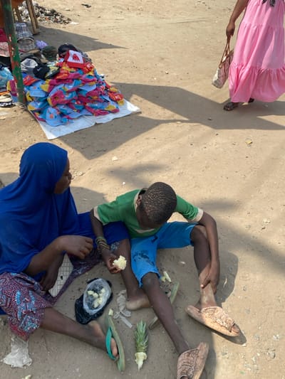 An outdoor market scene in Ablekuma, Ghana, features two individuals seated on an unpaved dirt surface in the foreground. A woman on the left, wearing a blue hijab and a patterned skirt, holds a piece of peeled pineapple. Next to her, a boy in a green t-shirt and blue shorts eats a piece of pineapple. A black plastic bag containing peeled pineapple chunks, a pineapple top, and discarded peels rest on the ground between them. The woman wears green flip-flops; the boy has one patterned slide on his right foot with the other slide lying adjacent. To the left, a large pile of colorful hats and apparel is displayed on a white sheet, positioned near a weathered wooden pole. In the upper right background, a person in a pink dress and sandals walks away, carrying a light-colored handbag. Multiple distinct shadows are cast across the sunlit ground.